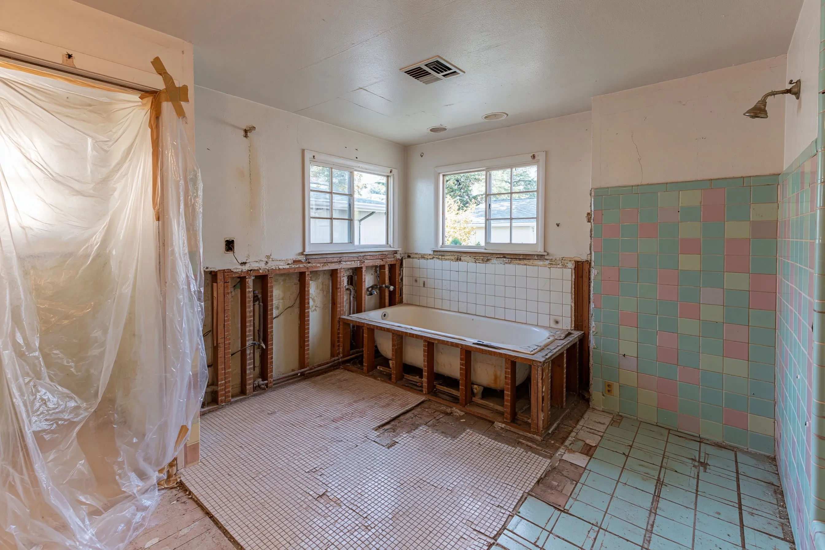Bathroom with the walls around an old built-in tub cut back to the studs, small floor tiles partly lifted, pink and green wall tile and plastic at the doorway